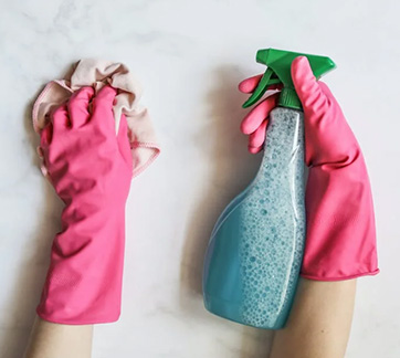 Two hands in pink rubber gloves, one holding a blue spray bottle and the other a beige cleaning cloth, against a white surface.